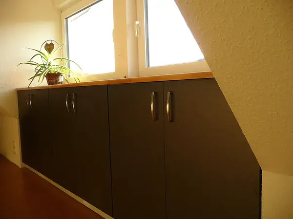 Sideboard unter Dachschräge mit dunklen Türen, Holzplatte und Zimmerpflanze am Fenster.