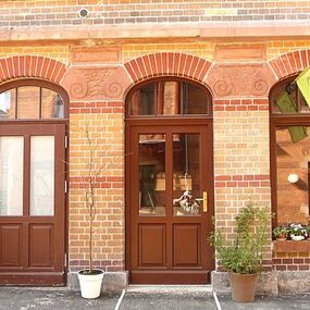 Eingangsbereich eines Altbaus mit braunen Türen, gelber Flagge und Blumen vor Backsteinfassade.